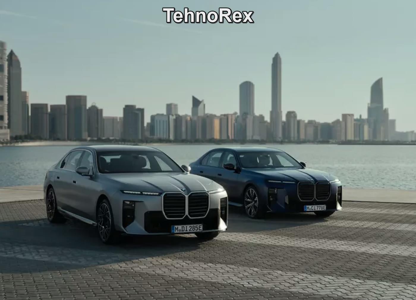 Два BMW i7, серебристый и синий, у воды на фоне небоскрёбов